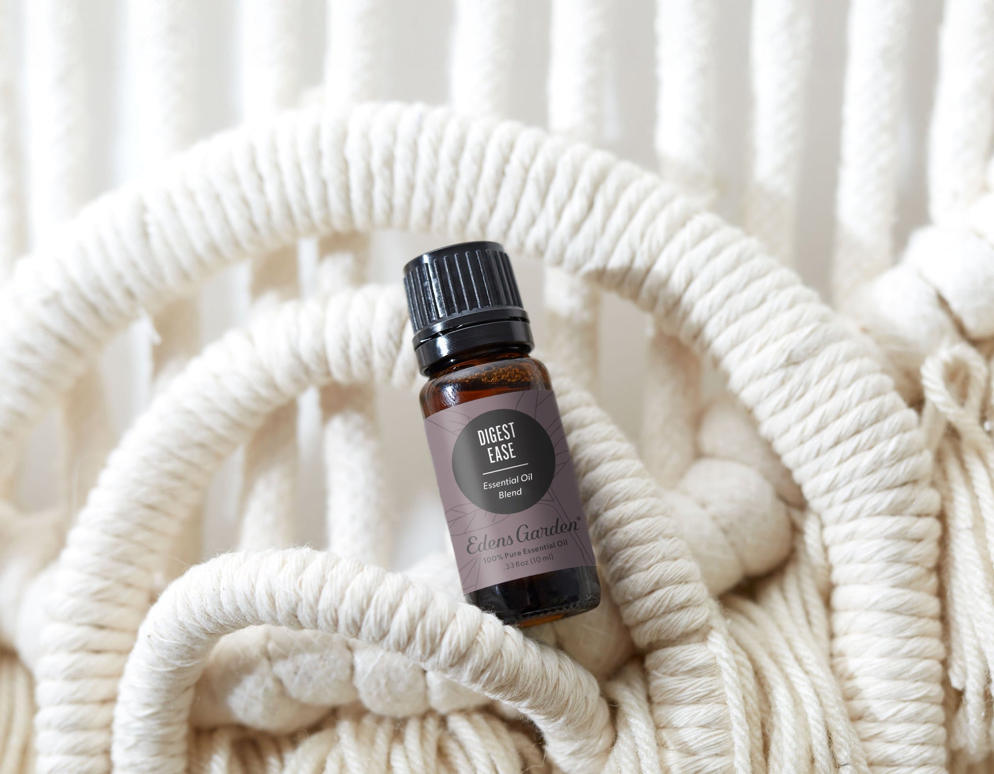 The image showcases a small amber glass bottle of “Digest Ease” essential oil blend by Eden’s Garden. The bottle is positioned centrally on a woven, circular placemat made of thick, off-white cotton rope with a neutral beige background.