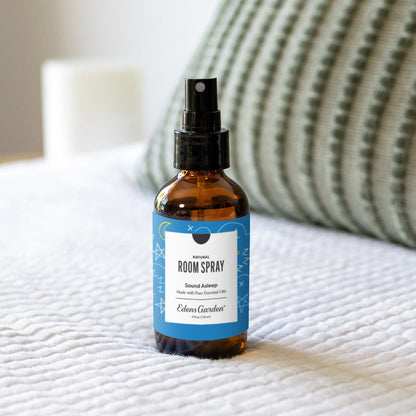 Sound Asleep ok for kids Room spray bottle on a white bed with a pale green textured pillow home setting with natural light coming in through the amber glass bottle and light shadow on the bed. 
