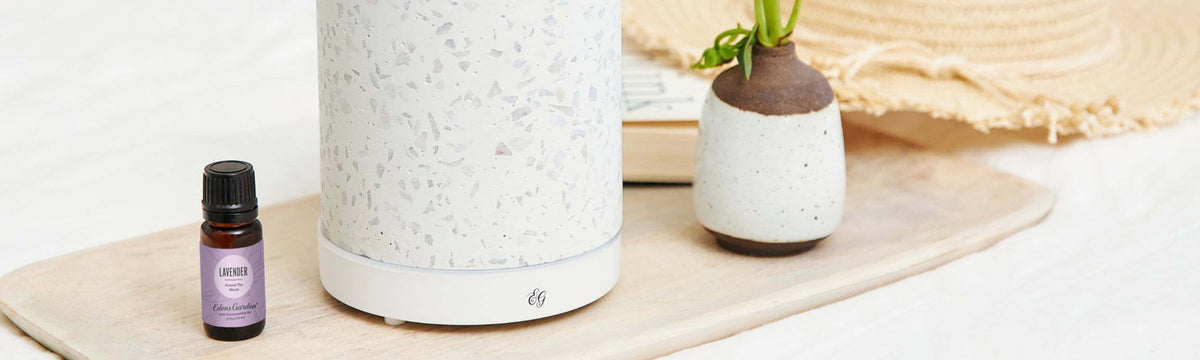 A bottle of essential oil and diffuser are on a wooden tray next to a small vase