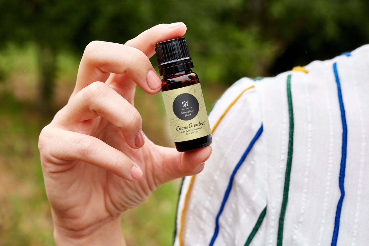 A woman wearing colorful strips on a white dress holds a bottle of Joy essential oil blend