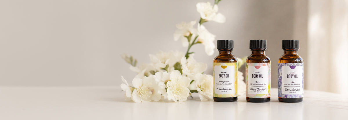 Three amber glass bottle body oils with black caps are lined up on a white surface next to white flowers against a soft beige background.