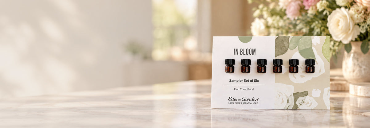 A boxed essential oil sampler set labeled 'IN BLOOM' stands on a light marble surface in a softly blurred indoor setting with flowers in the background. The package shows six small amber bottles and branding for Eden Garden.