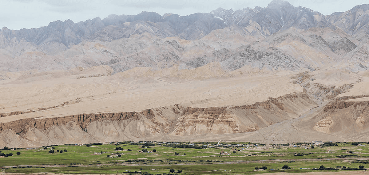 A wide landscape photograph of rugged tan and gray mountains rising behind a green valley with small scattered buildings and trees.