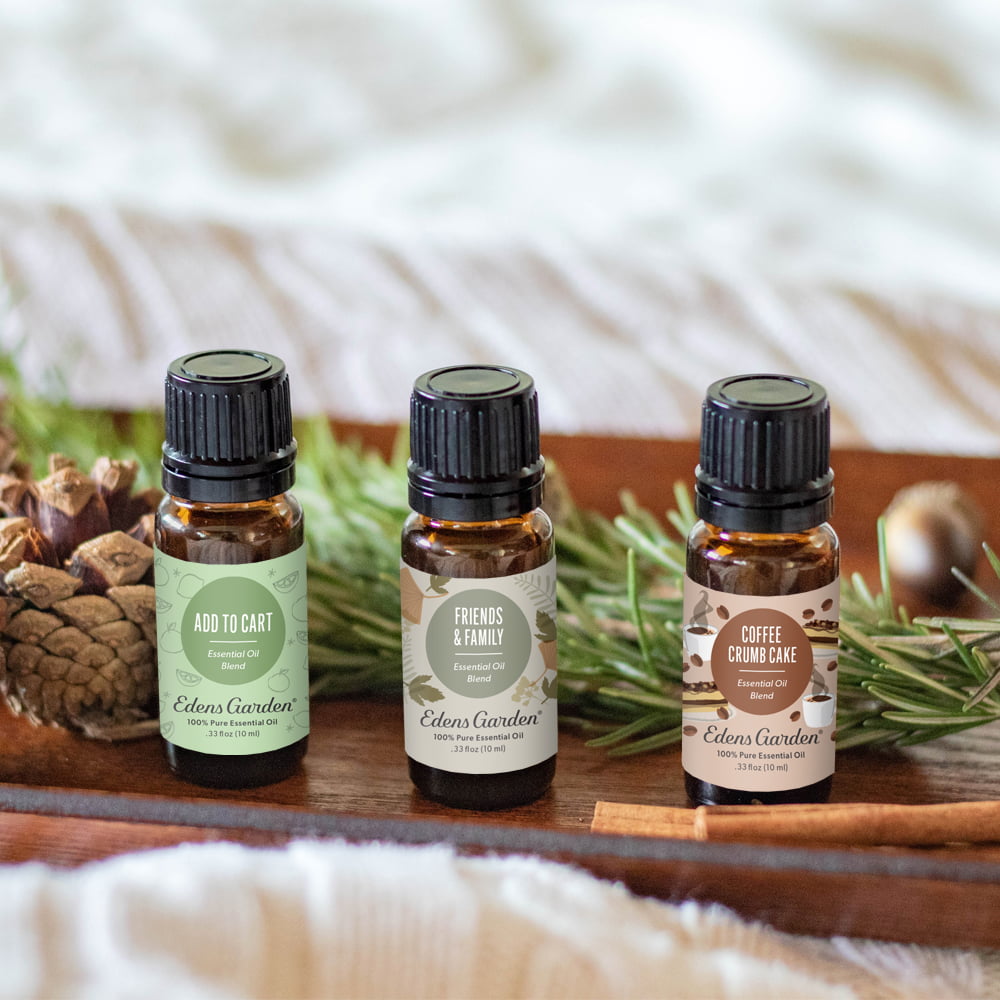 Three amber glass essential oil bottles with black caps from Edoss Gardens are arranged on a wooden tray with pinecones, rosemary, and a soft neutral fabric background. The labels read Add to Cart, Friends & Family, and Coffee Roasting.