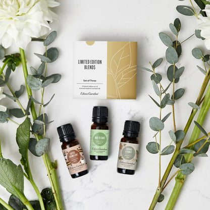 Three small amber essential oil bottles with black caps are arranged on a white marble surface with eucalyptus stems and white flowers around them. The bottles are part of a limited edition set from Edible Gardens, with the packaging box visible behind them.