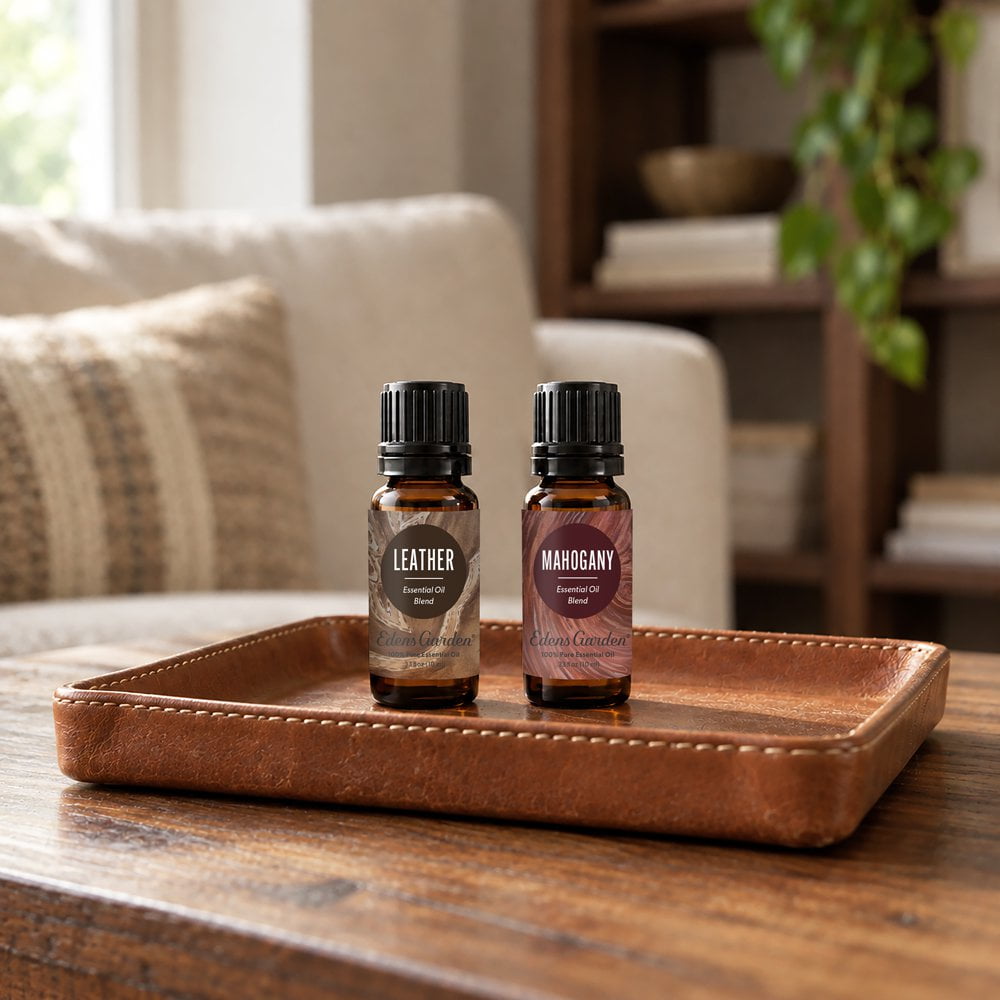 Two amber glass essential oil bottles with black caps sit on a brown leather tray on a wooden table in a softly blurred living room setting. The left bottle label reads LEATHER, and the right bottle label reads MARGAYNY.