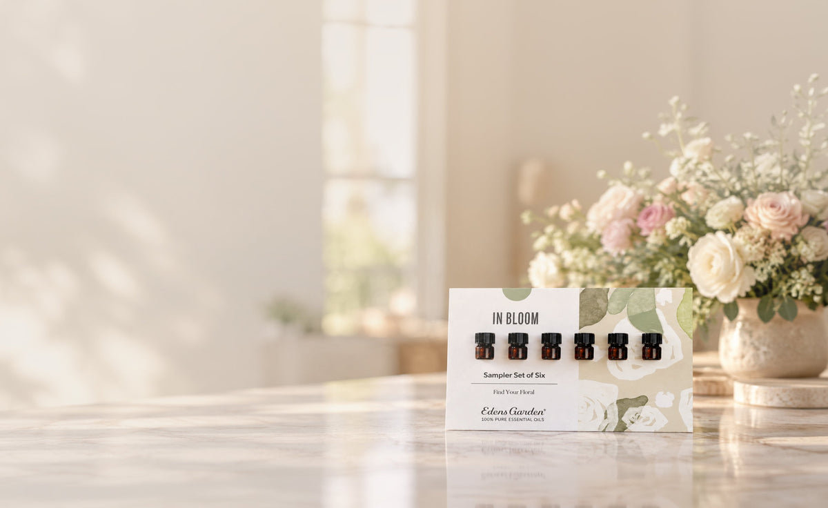 A boxed essential oil sampler set labeled 'IN BLOOM' stands on a light marble surface in a softly blurred indoor setting with flowers in the background. The package shows six small amber bottles and branding for Eden Garden.
