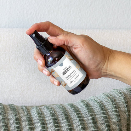 Hand holding an amber glass spray bottle labeled 'Sparkling Cinnamon Room Spray' against a neutral background.