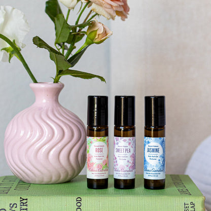 Three essential oil bottles labeled 'Rose', 'Sweet Pea', and 'Jasmine' on a book with a pink vase and flowers in the background.