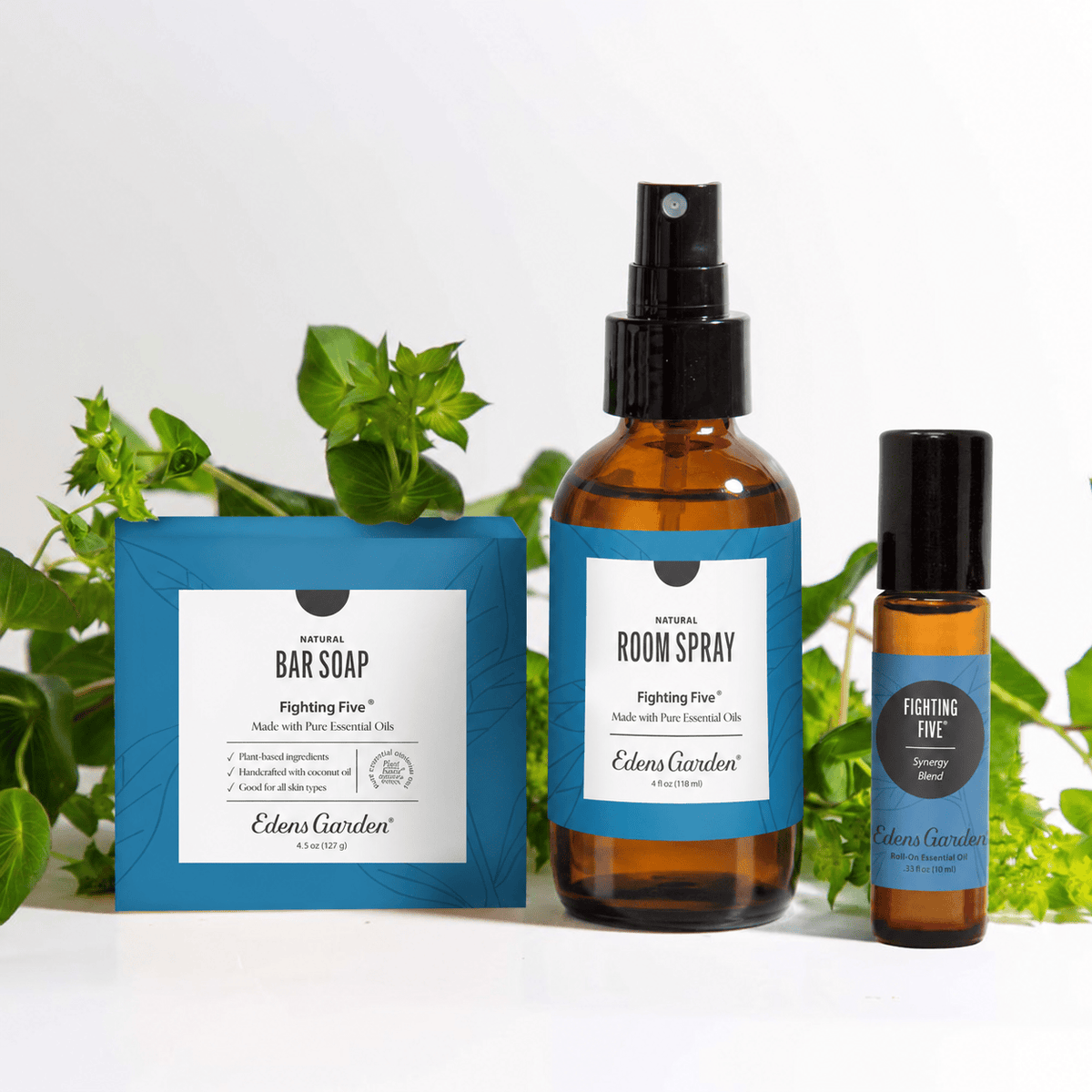 Three Edens Garden 'Fighting Five' natural products arranged on a white surface with green leaves behind them: a blue bar soap box, an amber room spray bottle, and a smaller amber oil bottle with a black cap.