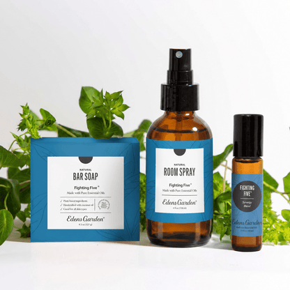 Three Edens Garden 'Fighting Five' natural products arranged on a white surface with green leaves behind them: a blue bar soap box, an amber room spray bottle, and a smaller amber oil bottle with a black cap.