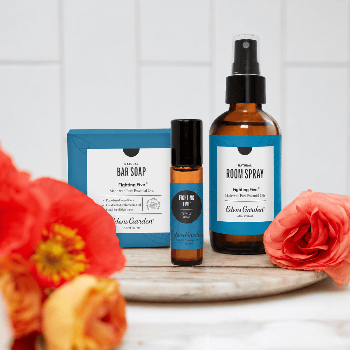Edens Garden essential oils room care set featuring Fighting Five spray, Fighting Five roll-on, and Fighting Bar Soap, arranged on a wooden board with blurred orange flowers in the foreground.
