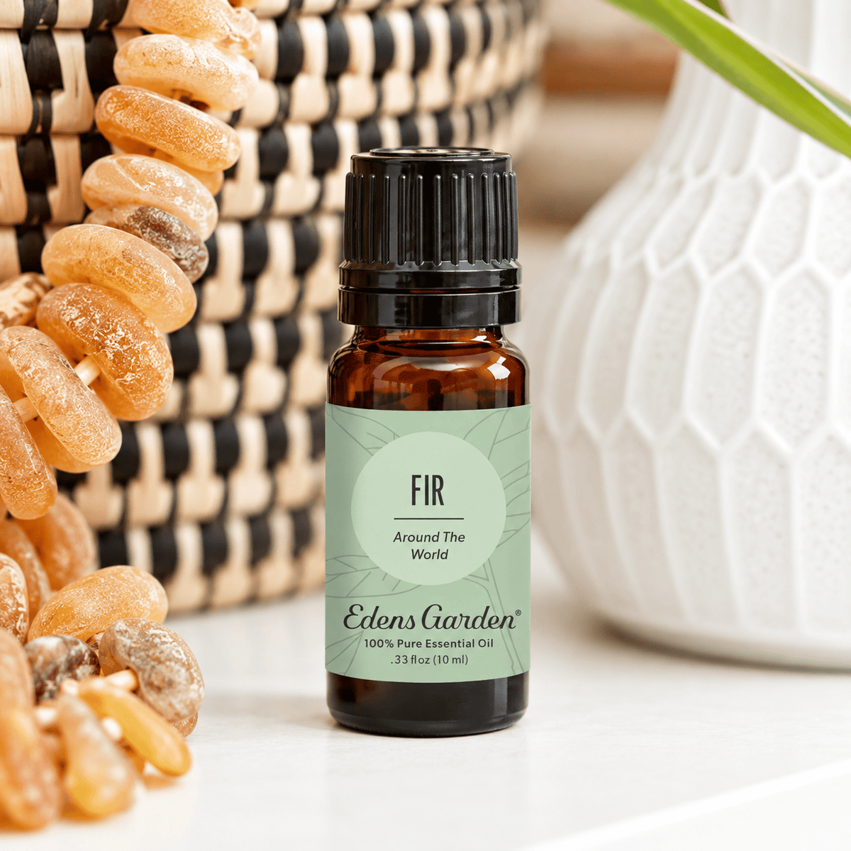 Edens Garden Fir Around The World 10 ml amber bottle on a white shelf beside woven baskets, amber beads and greenery.