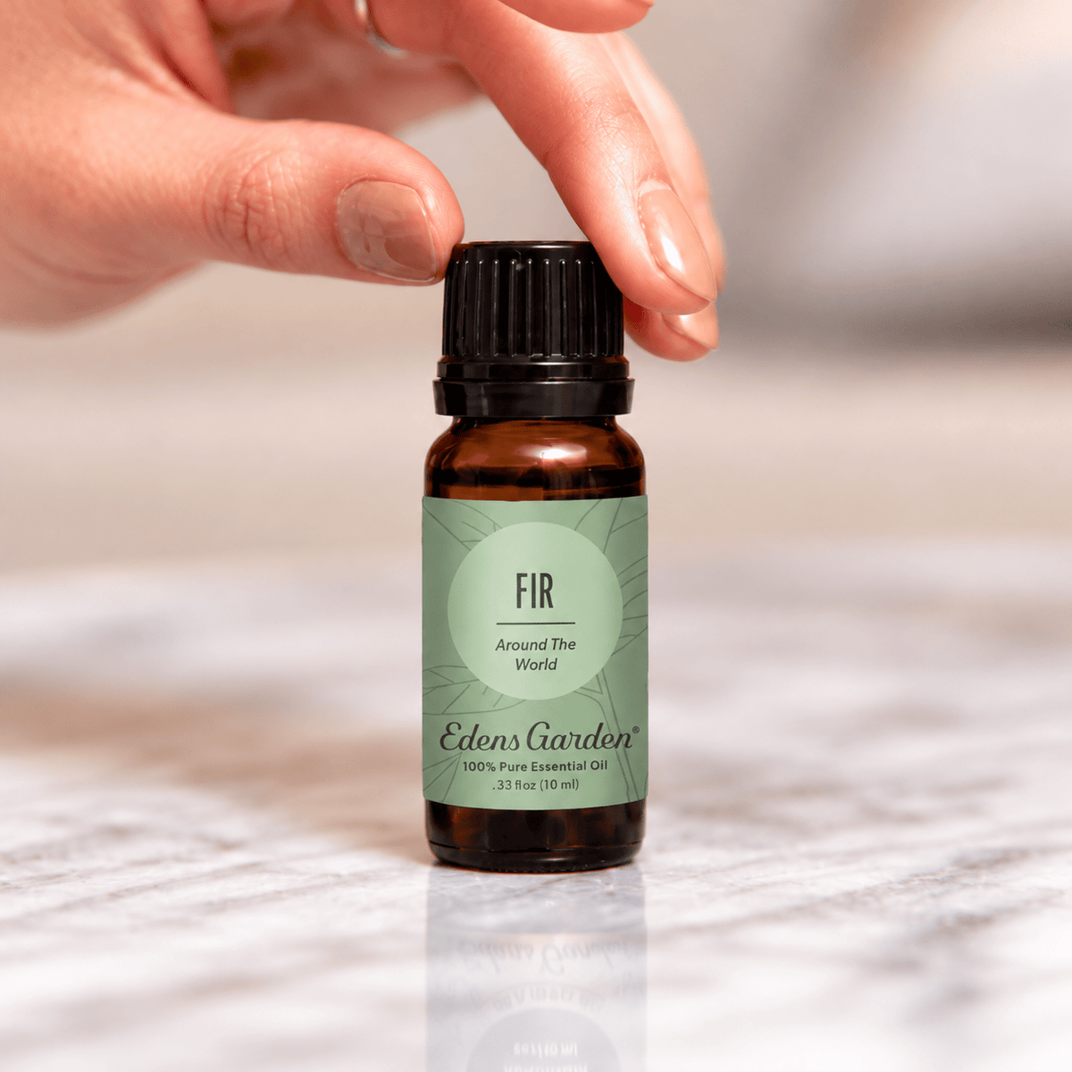 A hand reaching for an Edens Garden Fir Around The World 10 ml bottle on a marble surface.