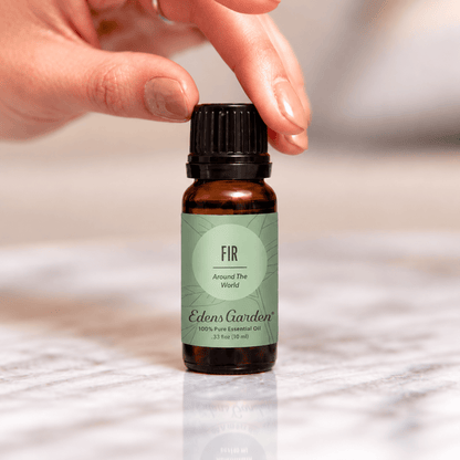 A hand reaching for an Edens Garden Fir Around The World 10 ml bottle on a marble surface.