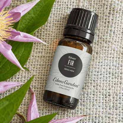 Edens Garden Silver Fir 10 ml amber bottle on textured linen beside purple flowers and green leaves.