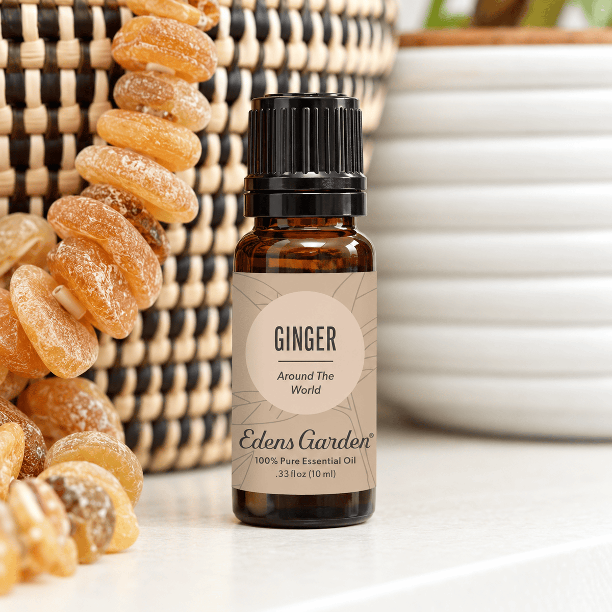 Edens Garden Ginger Around The World 10 ml amber bottle on a white shelf beside woven baskets, amber beads and greenery.
