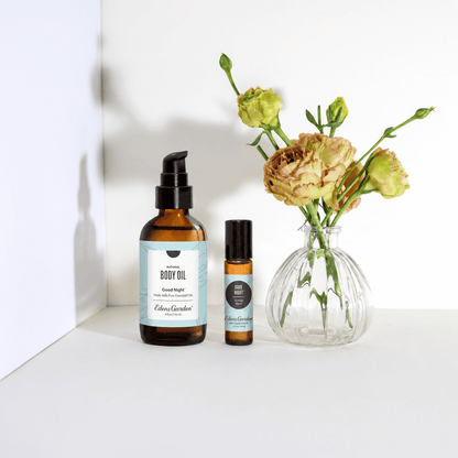Two amber glass body oil bottles with black pumps and light blue labels are displayed on a white surface next to a clear glass vase holding dried pale yellow flowers. The larger bottle is labeled 'Elixir Garden' and 'Good Night' body oil, and the smaller bottle is a matching rollerball.